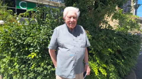 Keith Taylor, 84, with short white hair, wearing a pale blue shirt and beige chino trousers. He is standing in front of a bush on a sunny day and smiling at the camera.