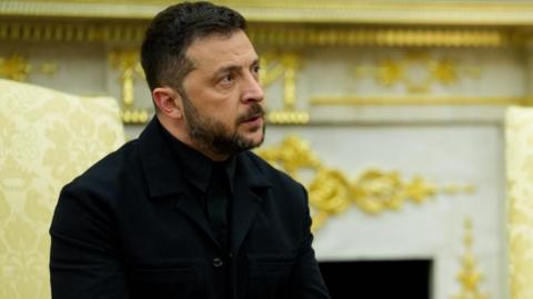 Volodymyr Zelensky sits in a cream-coloured chair. He's wearing a black jacket and black shirt.