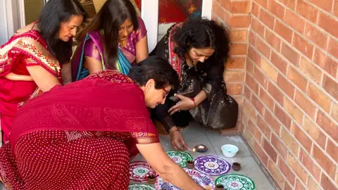 Four women in traditional hindu clothing squatting down lighting small oil-based candles