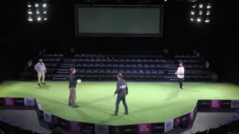 A stage has been turned into a rugby stadium with green carpet standing in for grass and seats for spectators. The cast are stood on stage with a rugby ball practising their lines and running through their positions.