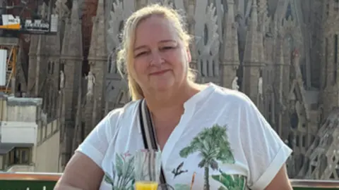 Alison has blonde hair in a ponytail, wears a white t-shirt with a palm tree design. She has a crossbody handbag on and holds a champagne flute with what appears to be orange juice inside. In the background is a large ornamental building which appears to be a cathedral.