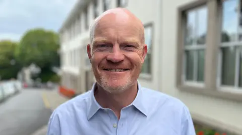 BBC Deputy Gavin St Pier, a bald man smiling at the camera, he has a grey patch on the beard and a ginger bit running up his face. He is wearing a blue shirt. 