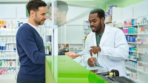 A stock photo of a male customer talking to a male pharmacist at a counter in a shop. Behind them both, on each side of the counter which has a screen, are shelves of medicines. The customer's face is reflected in the screen. 