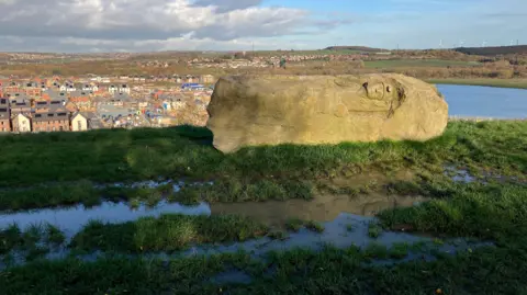 Chloe Aslett/BBC Taken from the top of a grassy, muddy hill, overlooking a big housing estate. There is a large rock, and in it, the face of a man is carved out, with him wearing a hat with a torch on.