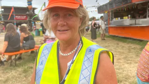 A woman is wearing a yellow high-vis vest and an orange cap. She is smiling. She is sitting in front of a food truck.