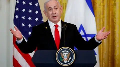 Benjamin Netanyahu, Israel's prime minister, speaks at a news conference. He is wearing a black suit with a red tie and is stood behind a lectern and in front of the flags of the US and Israel