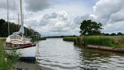 Owen Sennitt/BBC A yacht is moored up along the bank of a river on an overcast day. 