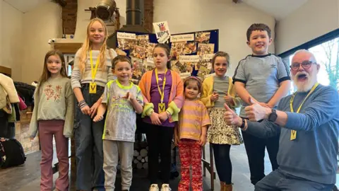 A group of six children of varying ages standing in what looks like a community hall. There is a man in a blue jumper and jeans, with a white beard kneeling down to the right.
