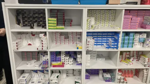 A white cabinet with about 16 cubbyholes each full of different medicines in packets piled high.