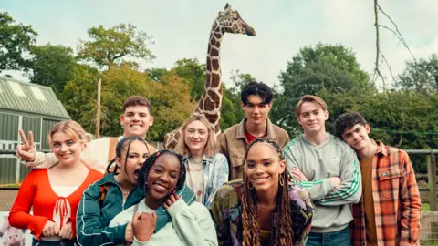 Netflix/Heartstopper Season 3 The cast of Heartstopper smile at the camera while filming on location at a zoo. A giraffe and trees are visible in the background.