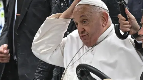 Pope Francis looks weary whilst riding in a popemobile on a trip to Belgium. He is dressed all in white.