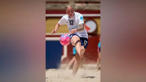 Jose Manuel Alvarez/Euro Beach Soccer A woman in a white football kick is facing the camera, kicking the ball with right foot. She is on a beach.