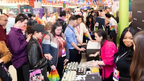 A busy room can be seen with a career fair taking place. Students can be seen talking to businesses at each stand 