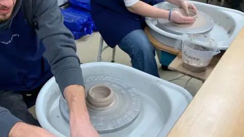Contributed A close up of two pottery wheel stations. A man and woman sit behind each as they work on clay.