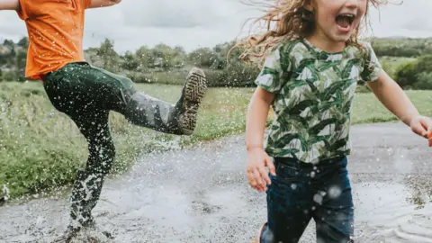 Two children joyfully play in a large puddle - a little girl runs as the older boy kicks water at her - stock photo