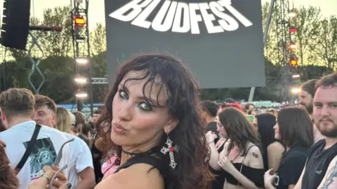 Sasha Sasha's got long curly brown hair and is pouting into the camera while in the crowd. She has cross earings and is wearing a black sleeveless top. She's in front of a big stage with a screen that says "Bludfest".