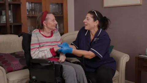 An older woman in a wheelchair, who is in her home, gets support from a Crossroads Care Surrey worker.