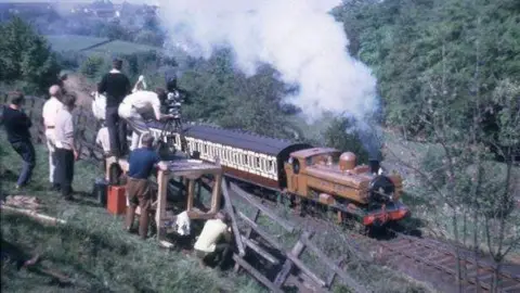 A mustard brown-coloured locomotive pulling two carriages with a film crew looking on from a hillside