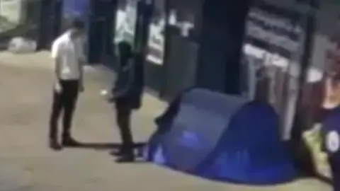 Gloucestershire Constabulary A man wearing a white t-shirt is seen talking to a man dressed in black clothing. They are standing next to a blue tent.