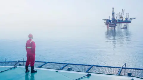 an oil worker in a red uniform and and hard hat stands on one North Sea platform looking across the water to another platform