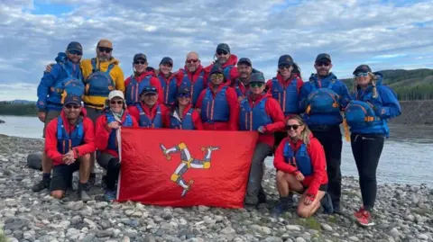 Expedition Limitless The Expedition Limitless group of 17 people wearing red coats and blue life jackets, hats and sunglasses. Half of the group is standing, the other half is kneeling in front holding a red Manx flag featuring the triskelion in white, grey and yellow. They are on a pebbled riverbank with hills in the background and a blue sky with white clouds above.