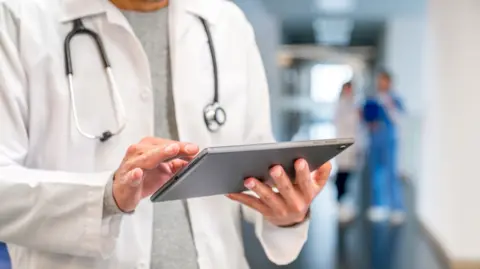 A doctor in a white medical coat reads from a tablet computer.