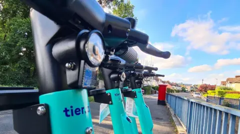 Cyan coloured e-scooters parked in a row on a residential street. In front of the scooters is a metal fence. Further down the road are houses and a car.