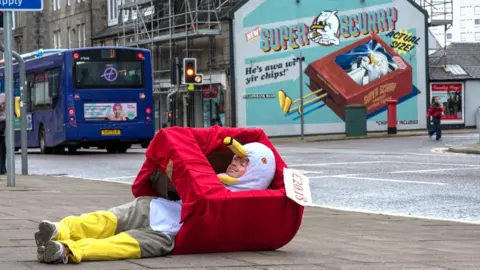 Man in a seagull costume, lying on the pavement, with an artwork of a seagull in the background on a wall that he is recreating.