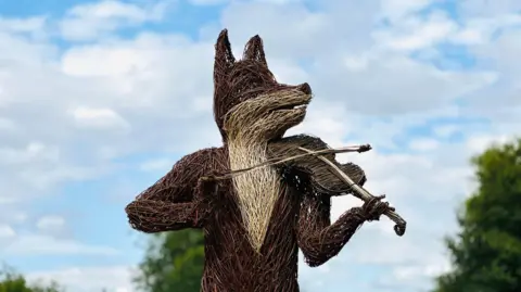 Hannah Olsson/BBC A large sculpture made out of twigs resembles a fox playing a fiddle. It stands in the field at the 2024 folk festival in Cambridge. It is a bright and dry sunny day with blue sky and a few white clouds. 