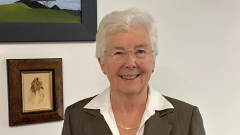 LDRS Ros Jones, mayor of Doncaster, is wearing a brown jacket over a white shirt. She has short grey hair and is wearing glasses