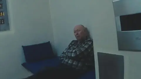 Stephen Alderton is wearing black trousers and a checked shirt, and is sitting in a police cell on a bed with a blue pillow and mat. There is the edge of a window on the left.