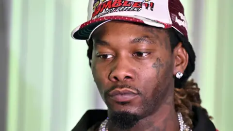 Offset is a man, he has brown eyes and is wearing a white and red cap. He has a diamond stud earring and is wearing a large silver chain. He is wearing a red jacket with a black collar. He has a tattoo on his cheek below his eye of a cross and tattoos on his neck. 