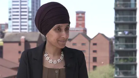 Sukhvinder Kaur is wearing a dark Sikh head covering or turban. He has a black jacket, with brown top and a striking necklace of white flowers. 