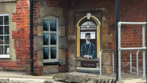 Nikita Spires How Nikita Spires' mural looked when she finished it, viewed on the wall of the station from a distance. On the left hand side, she has painted a window that looks like the others, complete with grey stones around it and six large panes. On the right hand side, she has painted an old-fashioned, Victorian ticket officer wearing a navy uniform and flat cap, sat waiting for tickets.