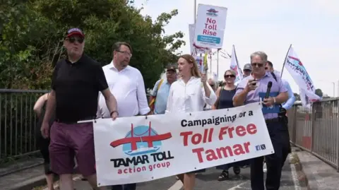 A group of people crossing the bridge. At the front are three men and one woman. They are holding picket signs and a banner which says Campaigning for a toll-free tamar. 
