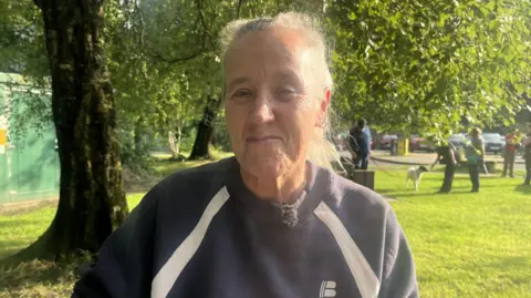 A woman standing in a park with a blue sweatshirt on. She has grey hair.