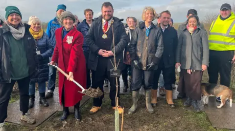 Nicola Haseler/BBC A group of about 16 people stand around the newly planted sapling, which has a protective plastic tube around it and a black label explaining what it is. The people wear winter coats and wellies or boots and smile at the camera. A lady wearing a red coat holds a silver and wooden spade with a pile of soil on it. 