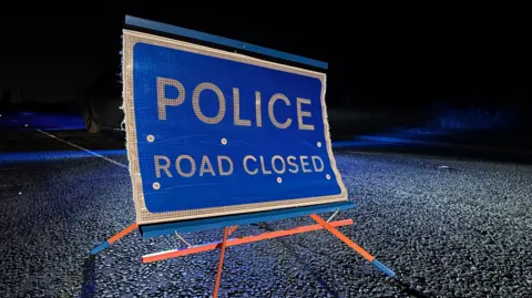 A blue sign with white writing that says POLICE ROAD CLOSED. It is set on an orange tripod on the road. 