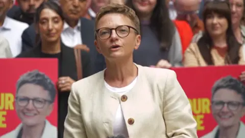 Bev Craig, wearing a white jacket, and in mid-speech. She has short blonde hair and is wearing glasses. There are people sitting behind her, as well as posters with her face on them.