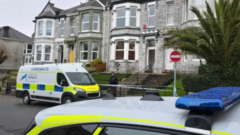 A police van and car outside a terraced property on Lipson Road in Plymouth. A police officer is standing guard at a cordon that is marked with police tape.