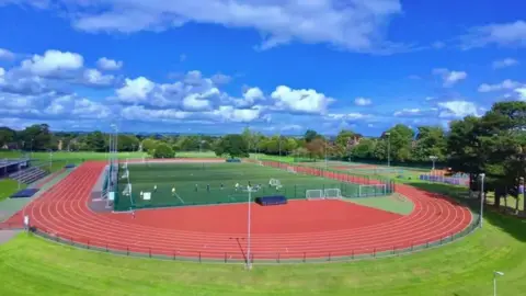 A green 3G football pitch in the middle of a red running track with stands to the left and tennis courts to the right. Trees can be seen in the distance under a blue sky.