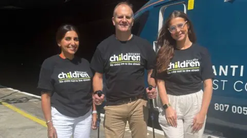 Two women and a man wearing black t-shirts with a white and green Railway Children writing and log on the front. They are standing on a station platform in front of a blue train. The woman on the left has black hair and white trousers and is smiling. The taller man has hiking poles in each hand and is wearing beige trousers. The woman on the right is younger, has long dark hair and transparent framed glasses. She is wearing beige trousers.
