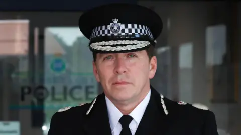 PCC Chief Constable Carden has short dark hair and blue eyes and is wearing a police uniform and looking into the camera. He is standing outside Merseyside Police building.