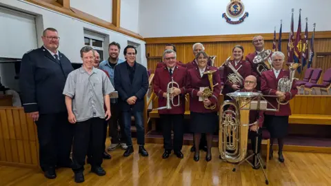 BBC Shakin' Stevens stood in the centre wearing a blazer and some jeans next to people from the charity and the brass band who are wearing red uniforms.