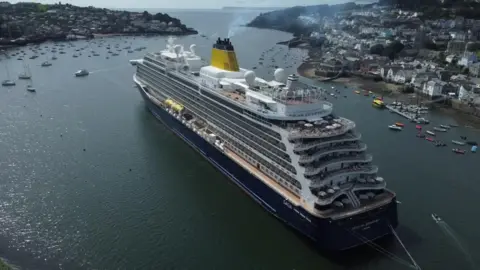 A very large cruise ship in the River Fowey, with smaller boats and land with houses on either side of it