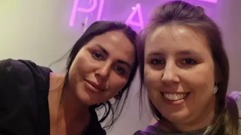 BBC Two wommen smile into camera. On the left is Alice who has black hair, and is wearing a black top. Her sister April it to the right. There is a neon sign behind them.