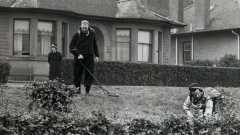 Getty Images Three police officers, two with metal detectors, are searching some ground and bushes outside a stone bungalow.