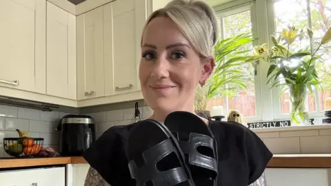 Emma Neil with blonde hair in a kitchen holding her sandals
