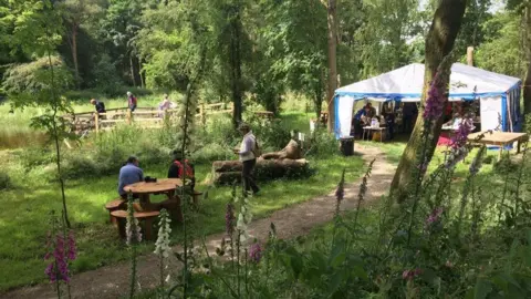 A path running through a woodland, leading to a white and blue tent. People can be seen walking or sitting on wooden benches. 