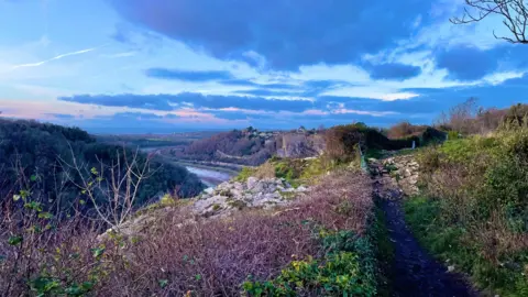 Fluffyclouds A blue sky with dramatic blue and orange clouds sits above the Avon Gorge. The trees and hedges are very sparse, with a little green visible among the branches and rocks. There is almost a purple light on the branches. The river, at very low tide, can be seen running through the gorge. 